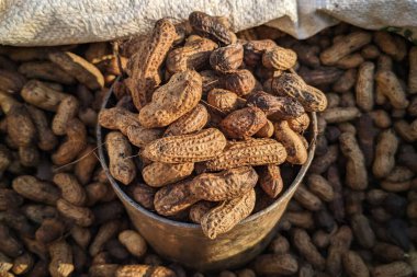 Kabuksuz fıstıklarla dolu metal bir kap. Fıstıkların kaba, desenli bir kabuğu ve kahverengi bir rengi var. Üs çevresinde daha fazla dağınıklık ve kırsal ve doğal bir görünüm oluşturarak üst üste yığılmış durumdalar..