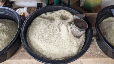 Pirinç yığınıyla dolu büyük siyah bir kap. Pirinç taneleri beyaz ve ince desenli. Konteynırda kısmen pirincin içine gömülü bir metal kepçesi var. Ölçmeye ya da satmaya hazır olduğunu gösteriyor..