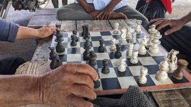 Tahta bir masada satranç oynayan bir grup insan. Satranç tahtasında iyi kullanılmış siyah ve beyaz parçalar var. Oyuncular, elleri tahtada hazır bir şekilde odaklandılar..
