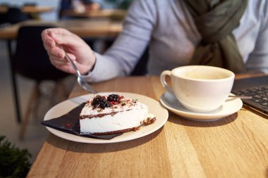 Kafede bir parça krem peynirli tatlı ve arka planda dizüstü bilgisayarla çalışan bir kadın.
