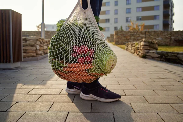Person holding a string bag with fresh vegetables and fruits in the street