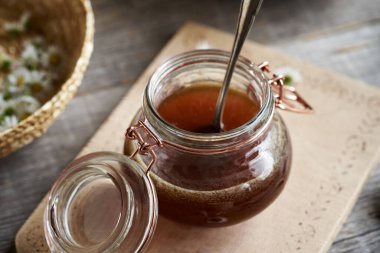 A jar of herbal syrup with a basket of common daisy flowers in the background clipart