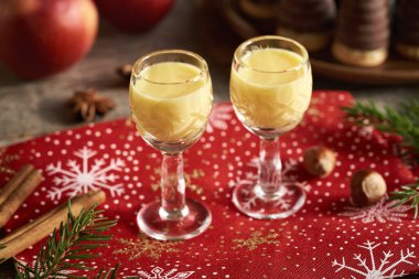 Arka planda elma ve Noel kurabiyeleri olan iki küçük bardakta eggnog.