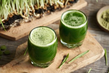 İki cam fincanda arpa otu suyu, arka planda taze bıçaklar yetişiyor.