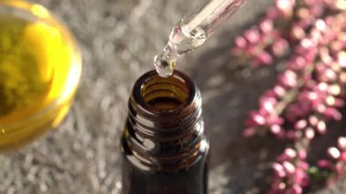 Aromaterapi damlaları bir damlacıktan kahverengi cam bir şişeye dökülür ve arka planda taze pembe çiçekler vardır.