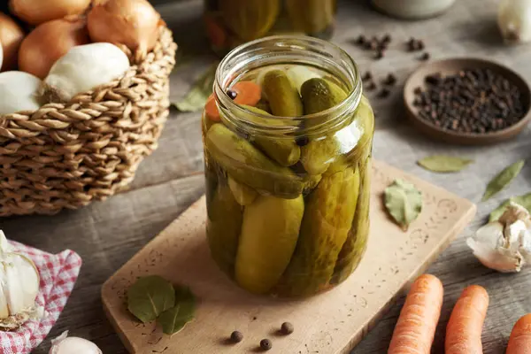 Homemade cucumber pickles in a glass jar on a table with carrots, onions and spices