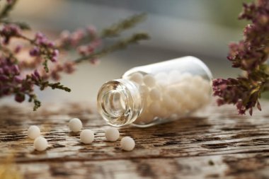 Bir şişe beyaz homeopatik hap ya da küre pembe kurumuş çiçeklerle dolu bir masanın üzerine dökülmüş.