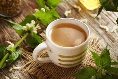 Bir fincan bitkisel çay, beyaz ölü ısırgan otu çiçekleri ve masada yapraklar.