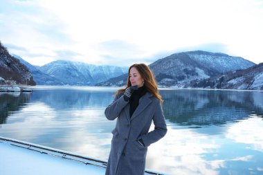 A serene woman in a coat and mittens enjoys the peaceful snowy fjord and mountain views in Gaupne, Norway, during winter.  clipart