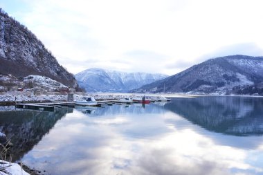 Norveç 'in Gaupne şehrinde sakin sular, karla kaplı çevreler ve çarpıcı dağ yansımaları ile kenetlenmiş teknelerin bulunduğu huzurlu bir marina.