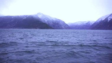 Suyun üzerinde dalgalanan karla kaplı dağlarla çevrili sakin bir kış gölü, bulutlu bir gökyüzünün altında Norveç fiyordunun manzarasında geçiyor.. 