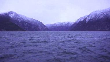 Soğuk dalgalı su, karla kaplı dağlar ve bulutlu bir gökyüzünün altında sakin bir atmosfer içeren kışın huzurlu bir Norveç fiyordu.. 