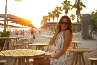 Huzurlu bir sahil kafesinde güneş gözlüklü şık bir kadın gün batımının tadını çıkarıyor. Tahta mobilyalar, kumlu yollar ve palmiye ağaçlarıyla çevrili sıcak altın ışık, rahatlatıcı bir tatil sahnesi yaratır.. 