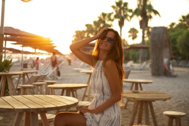 A smiling woman in a summer dress and sunglasses sits at a wooden table at a beachside cafe during sunset. The warm golden light, palm trees, and serene surroundings create a peaceful vibe.  clipart