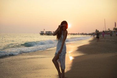 Gün batımında dalgalı bir elbiseyle yürüyen, sakin dalgalar, yumuşak kum ve sıcak altın ışıkta yıkanmış, huzur ve doğal güzelliği çağrıştıran pitoresk bir iskeleden oluşan sakin bir sahil sahnesi. 