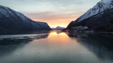 Altın bir kış günbatımı, Norveç fiyortlarının üzerine ılık bir parıltı saçar, sakin karlı dağlar ve sakin bir güzellikle çevrelenir.. 