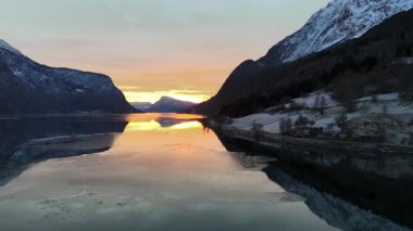 Altın bir kış günbatımı, sakin bir Norveç fiyordunu aydınlatır. Karlı dağlar ve sakin yansımalarla insansız hava aracının bakışından huzur veren bir güzellik yaratır..