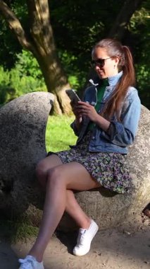 Parkta güneşli bir günün tadını çıkaran güzel bir kız telefonuna bakarken gülümsüyor. Rahatlamış ve neşeli, yemyeşil yemyeşil ve huzurlu bir ortamda günün sıcaklığını kucaklıyor..