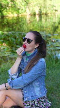 Parktaki yeşil çimlerde oturan bir kız güneşli bir yaz gününde taze çilek yiyor. Bereketli yeşillik ve ılık güneş ışığıyla çevrelenmiş, gevşiyor ve huzurlu ve neşeli atmosferin tadını çıkarıyor..