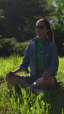 Güzel bir kız güneşli bir günde yeşil çimlerde huzur içinde meditasyon yapar. Yakın plan, canlı yeşillik ve güneş ışığıyla çevrili sakin ifadesini yakalar..