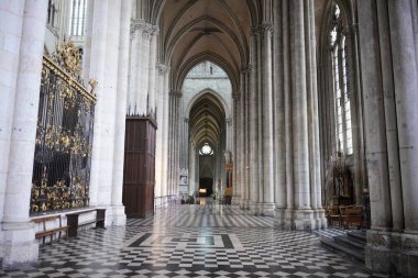 Yükselen kemerler, siyah-beyaz kareli bir zemin ve gün ışığıyla aydınlatılan süslü demir işlerinin yer aldığı uzun bir Gotik katedral koridoru..