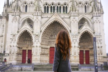 Fransa Amiens 'teki büyük Gotik katedrale bakan gri paltolu, uzun saçlı bir kadının arka planında süslü taş detaylar ve kırmızı kapılar var.. 