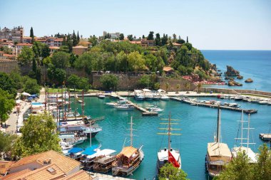 Geleneksel ahşap tekneler, turkuaz sular, tarihi taş duvarlar, bereketli yeşillikler ve güneşli bir gökyüzü altında manzaralı bir sahil manzarası olan Antalya marinasının çarpıcı manzarası.. 
