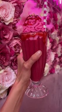 Pamuk helva, krem şanti, renkli süsler ve şurup ile kaplanmış canlı pembe bir milkshake. Çiçek arkaplanının önünde duruyor.. 