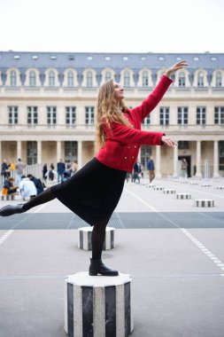 Kırmızı ceketli ve siyah elbiseli şık bir kadın Paris, Palais-Royal 'de çizgili bir sütunda zarif bir bale pozu veriyor. Tarihi mimari ve çevreleyen insanlar sanatsal açıdan gelişiyor..