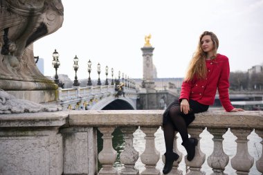 Kırmızı paltolu şık bir kadın Paris 'teki 3. Alexandre Köprüsü' nün taş korkuluklarında zarif bir şekilde oturuyor. Süslü köprü, altın heykeller ve klasik sokak lambaları romantik manzarayı güzelleştirir.. 