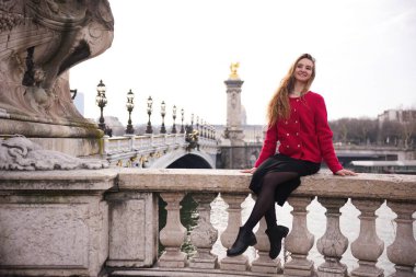 Neşeli bir kadın Paris 'teki 3. Alexandre Köprüsü' nün kayalıklarında oturmuş manzaranın tadını çıkarıyor. Tarihi köprü, süslemeli lambalar ve altın heykeller klasik bir Fransız arka planı sağlar..