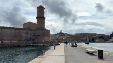 Marsilya, Fransa 04.14.2025 Rıhtımın yakınındaki müstahkem duvarlarda duran tarihi bir deniz feneri manzaralı bir liman geçidi ve uzak bir bazilika.