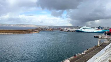 Marsilya, Fransa 04.14.2025. Sanayi binaları, dalgakıranlar ve bulutlu bir gökyüzünün altındaki uzak dağlarla çevrili büyük feribotları olan yoğun bir liman. 