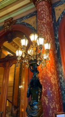 03.18.2025 Monaco, Fransa, Zarif Monte-Carlo iç mimarisi kristal bir avize tutan bronz bir heykel sergiliyor, mermer kolonlar, karmaşık ahşap işçiliği ve sıcak ortam aydınlatması ile çevrili..