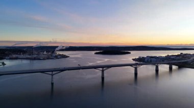 Gündoğumunda İsveç, Sundsvall 'ın hava manzarası, modern bir köprü, dumanlı endüstriyel liman ve sakin sabah sularına hafifçe yansıyan altın bir gökyüzü..