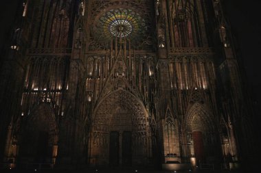 Strasbourg Katedrali 'nin simetrik gece görüntüsü Gotik ön cephe, aydınlatılmış gül penceresi ve üç süslü giriş kapısı. 