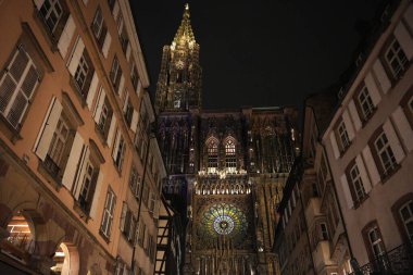 Strasbourg Katedrali 'nin nefes kesici gece manzarası süslü faşat ve parlayan gül penceresi, çevresindeki geleneksel binalar tarafından çerçevelenmiş.