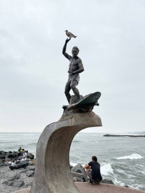 Lima. Peru 12.10.2024 Kayalık sahil boyunca dalgalar üzerinde sörf yapan bronz bir heykel duruyor. Heykellerin üzerine tünemiş bir kuş ve insanlar aşağıdaki kıyıda rahatlıyor.. 
