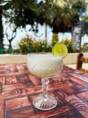 Soğuk bir Pisco Sour, tropik çiçekler ve palmiye ağaçlarıyla çevrili, açık bir kafedeki renkli bir masada taze limon dilimli eski bir bardakta servis edilir..