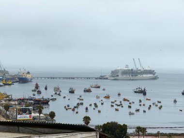 Chimbote, Peru 11.26.2024 Sakin bir sahil gününde, bulutlu bir gökyüzünün altında balıkçı tekneleri ve kargo gemileriyle dolu kalabalık bir limana büyük bir yolcu gemisi yanaşıyor..