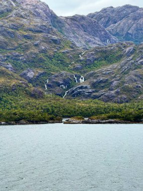 Çok sayıda küçük şelale taşar kayalık bir dağ sırasının yeşil orman yamaçlarında bulutlu bir gökyüzünün altında sakin bir fiyorda akar..