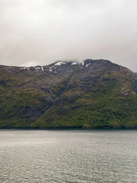 Yeşil yamaçları ve dağınık kar yamaçları olan sislerle kaplı bir dağ sakin bir fiyort üzerinde yükselir ve dingin ve karamsar bir doğal manzara yaratır.. 
