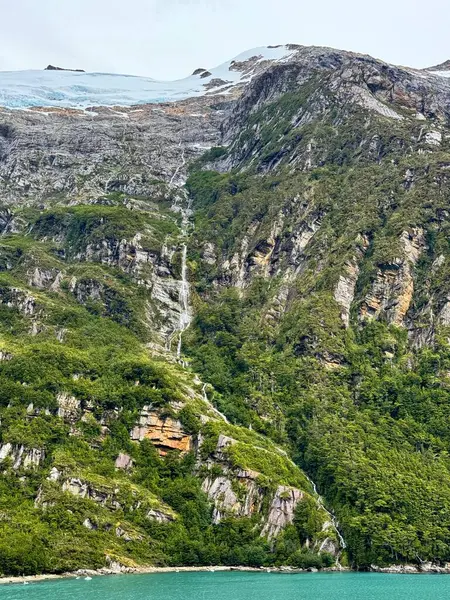 Dar bir şelale, karla kaplı bir buzuldan, sarp kayalıklardan, sık yeşil ormanlara, oradan da aşağıdaki parlak turkuaz fiyorda akar..