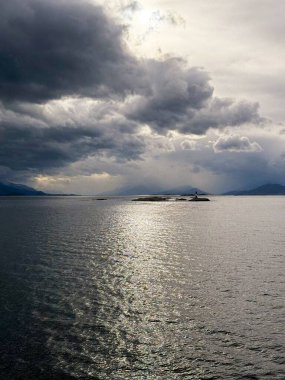 Karamsar bulutların altında, filtrelenmiş güneş ışığını yansıtan parıldayan suyla uzak kayalık bir adada bir deniz feneri duruyor..
