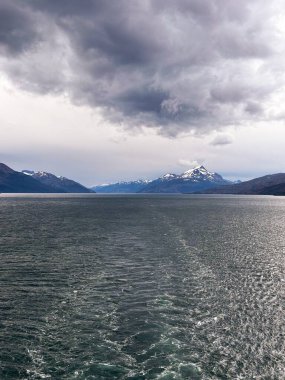 Kara bulutlar dramatik bir fiyortta beliriyor. Karla kaplı tepeler ve dalgalı sular bir gemi yolundan geliyor..
