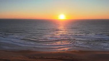 Yumuşak dalgalar ve dokulu kumlu plajlar üzerine yumuşak yansımalarla okyanusun üzerinde altın gün batımını gösteren, huzurlu sahil ışıklarını ve doğal güzellikleri sıcacık ve sakin deniz manzarasında yakalayan drone görüntüsü..