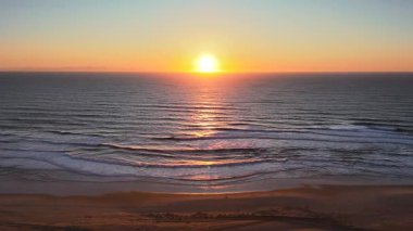 Sakin okyanus dalgaları üzerinde gün batımının hava drone çekimi, sıcak akşam ufkunda yumuşak sörf ve dokulu plaj kumları ile altın ışıkta parlayan. Huzurlu ve manzaralı bir sahil anı