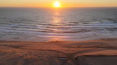 Okyanus dalgaları üzerinde altın gün batımı plaj yapıları ve hava aracı tarafından yakalanan yumuşak yansımalarla doğal güzelliği ılık akşam ışığında ince bir kıyı gelişimiyle harmanlıyor.. 