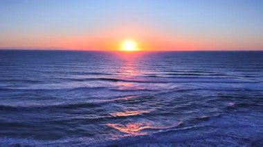 Okyanus dalgaları üzerinde renkli günbatımının görüntüsü, parlayan pastel yansımaları ve sakin ve manzaralı bir sahil şeridi boyunca yürüyen insanlar.. 