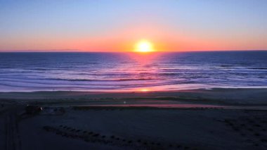 Dalgaların yumuşak pastel yansımalarıyla okyanusun üzerinde canlı günbatımının videosu ve kıyı şeridi boyunca huzur içinde yürüyen insanlar, sakin ve renkli bir kıyı akşamı yakalıyorlar..